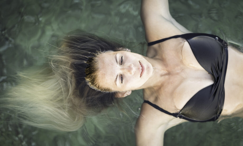 Eine junge, hübsche Frau liegt mit geschlossenen Augen und auf die Seite gestreckten Armen auf einer grünen Wasseroberfläche.