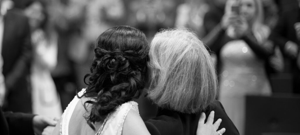 The bride and her best friend from the back view. Both put an arm around each other's shoulder and nestle together. In the background the wedding guests photograph this scenario.