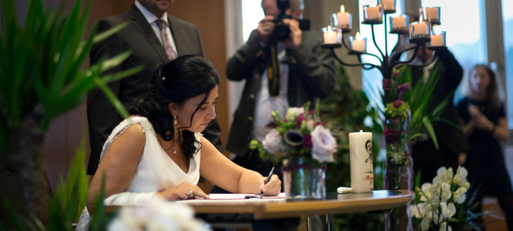 The bride signs the official documents at the registry office. In the background photographers and wedding guests.