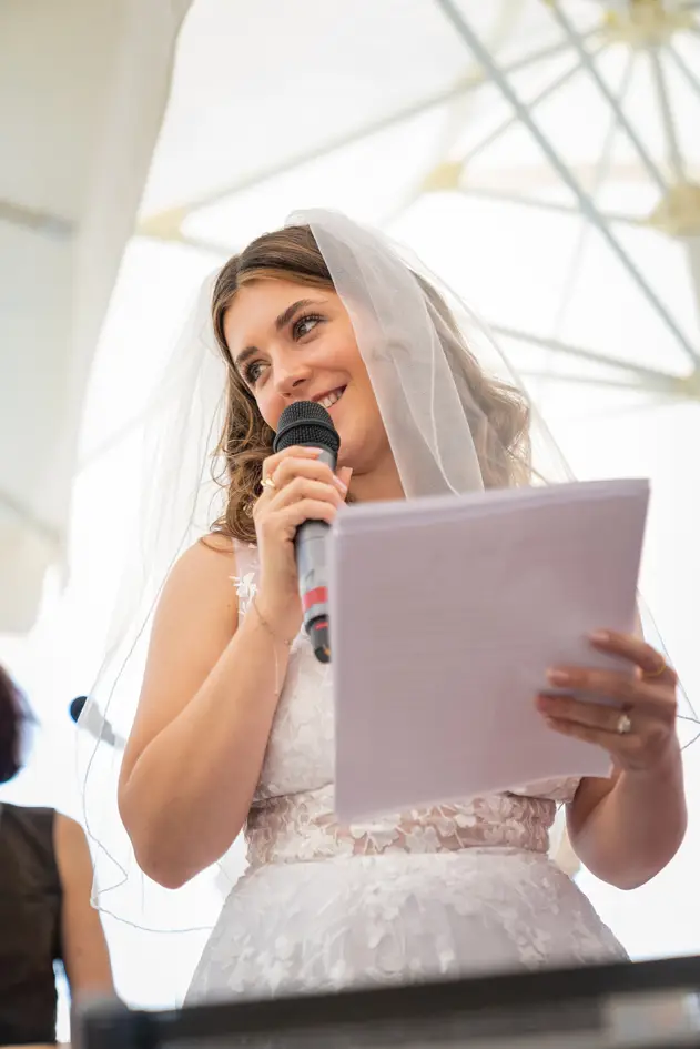 The bride gives her wedding speech. In one hand she holds a microphone, in the other a large sheet of paper. She smiles charmingly as she does so. In the background, a sea of white parasols.