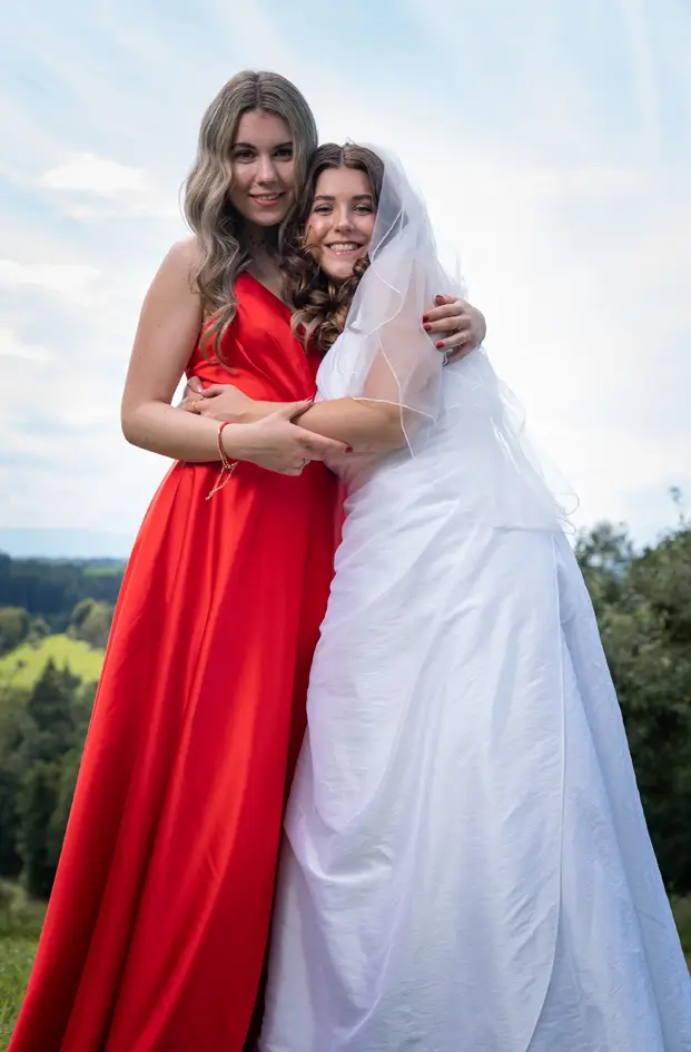 The bride in her beautiful wedding dress lovingly and smilingly snuggles up to her friend who is wearing a charming red dress.