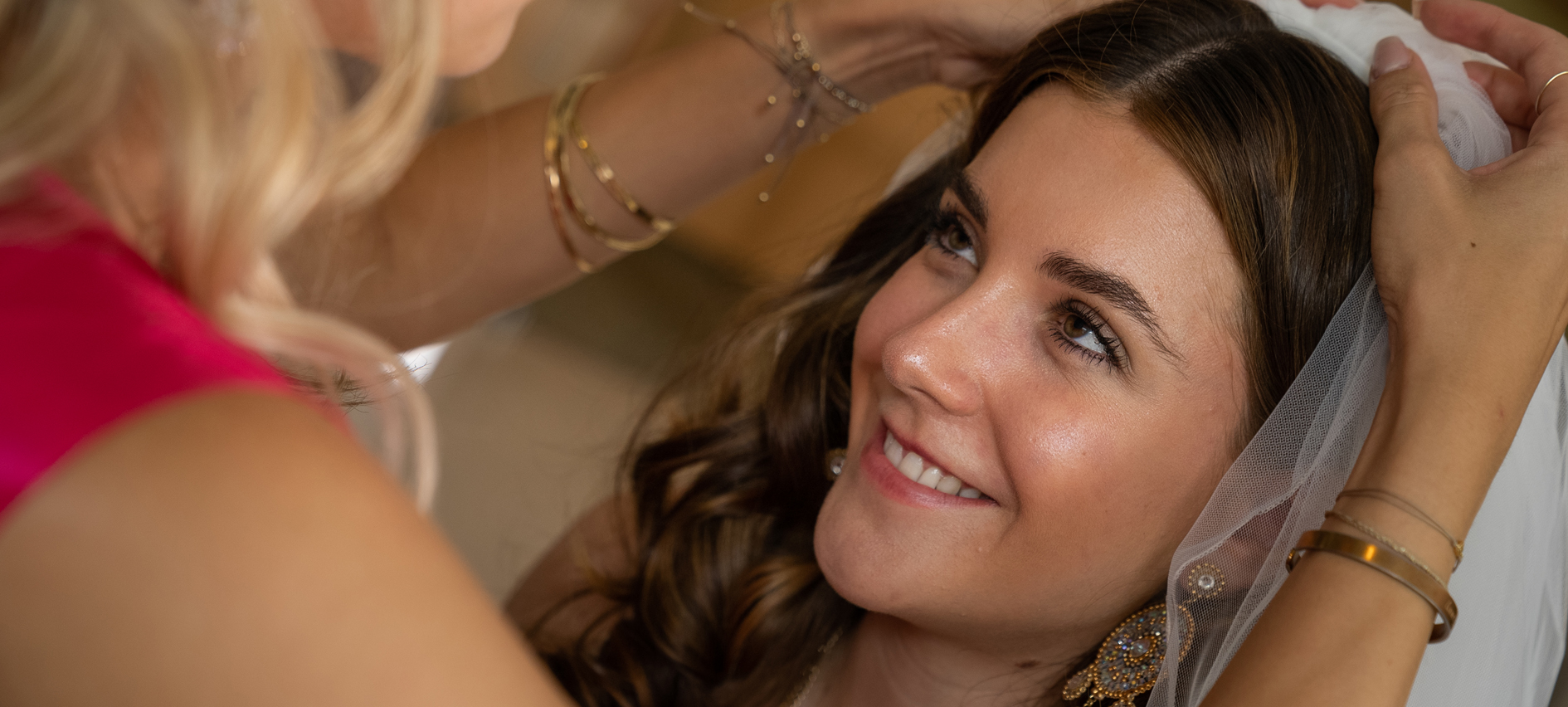 A friend puts the bridal veil on the bride's hair. The bride gives her a radiant smile.