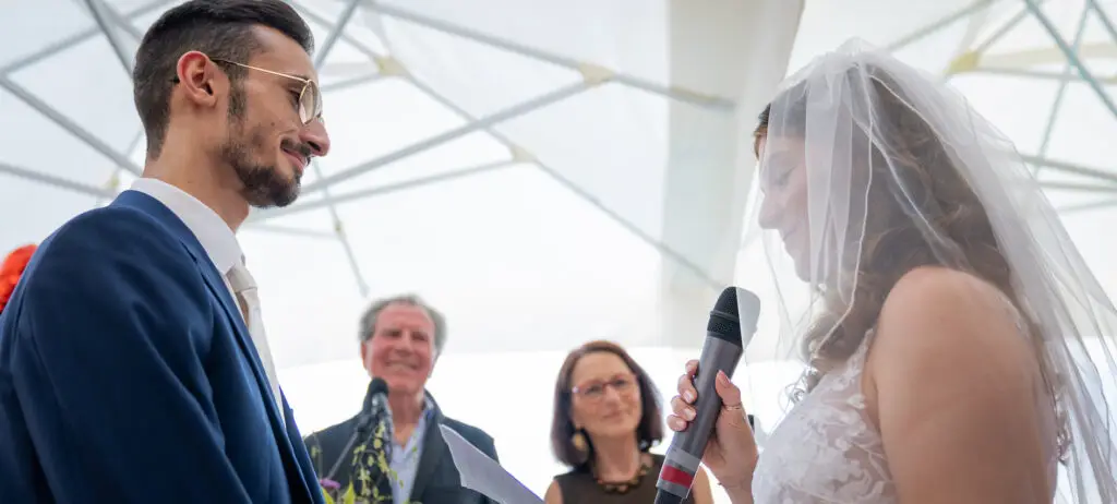 The bride holds a microphone in one hand and a piece of paper in the other and gives her speech to the groom and her family.