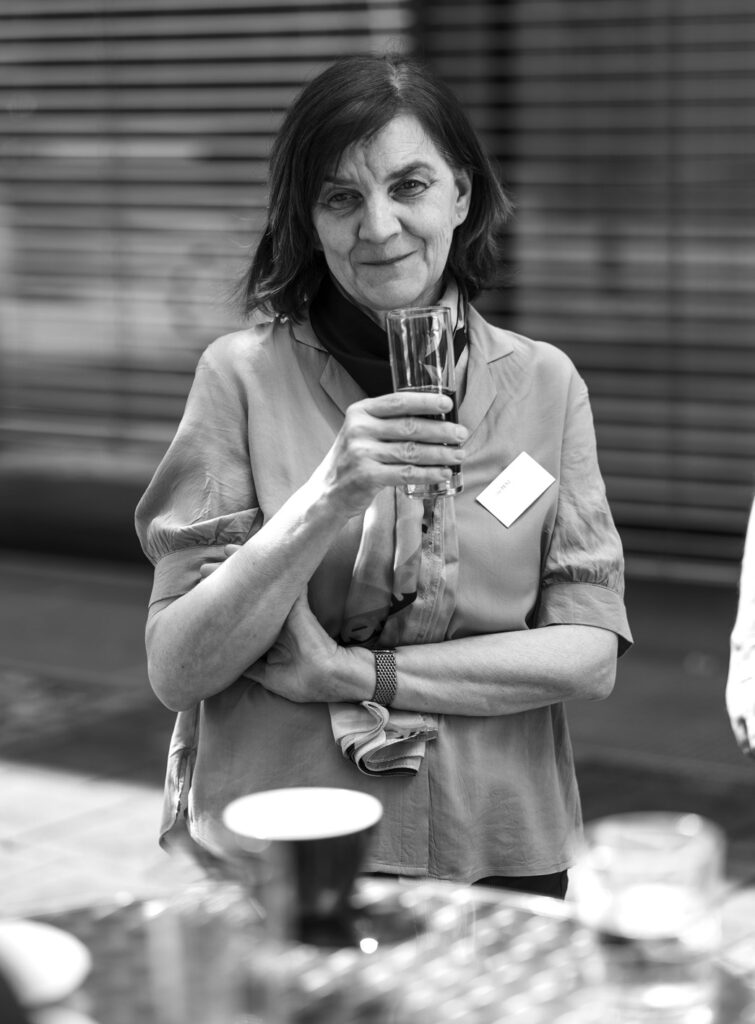 A lady about 50 years old, holding a glass of water in her hand, looks benevolently into the camera. In the background lowered blinds.
