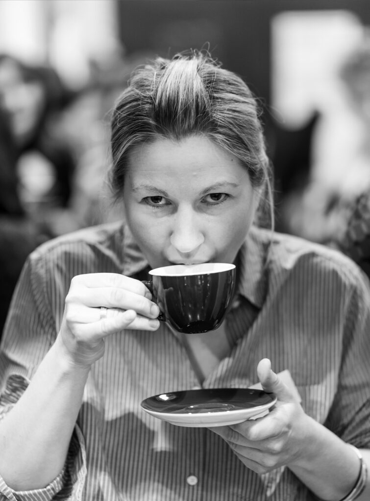 A lady about 30 years old brings her teacup to her mouth to drink, in the other hand she holds the saucer. She looks toward the camera with interest.