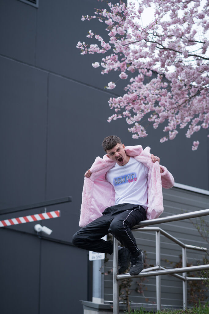 The young rapper sits on a silver outdoor stair railing wearing a plush pink jacket. He tears the two jacket parts apart with his hands. In the background a tree blooming with pink flowers.