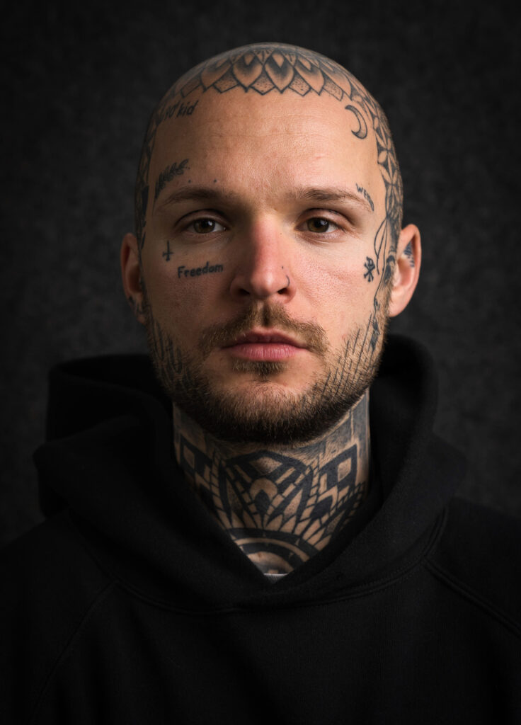 Portrait photography of the tattoo artist. He looks into the camera and shows his art painting applied to the skin on the head and neck.