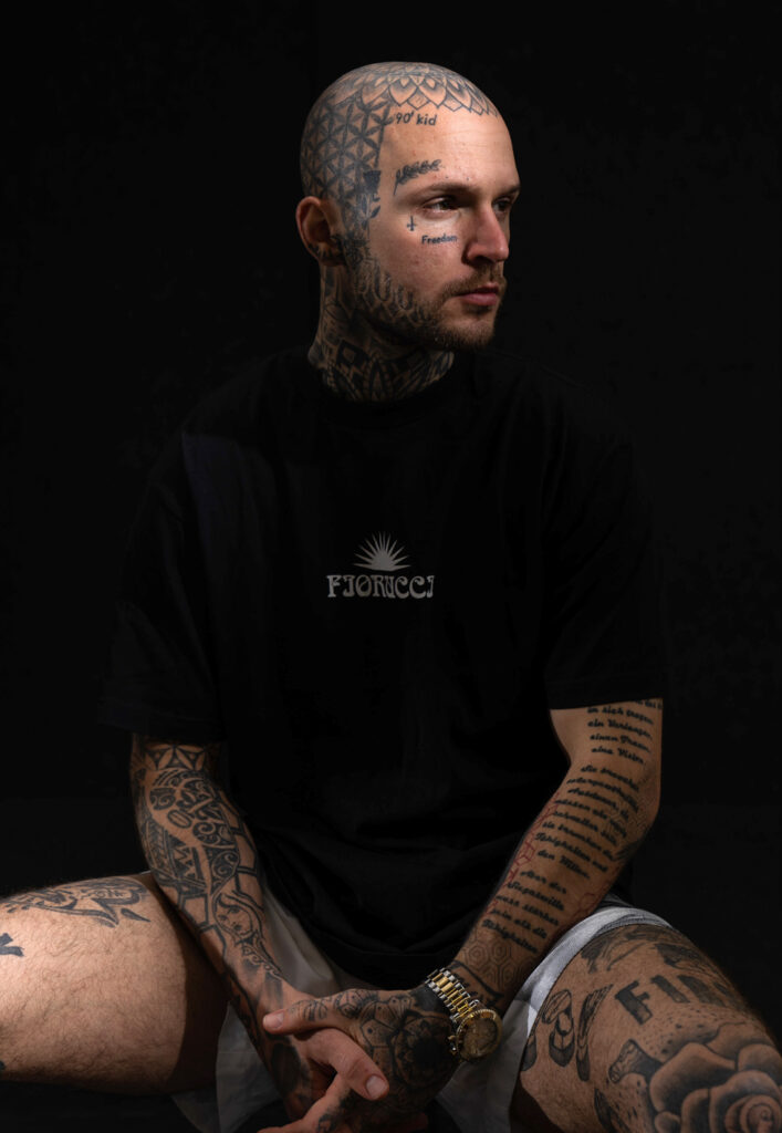 The young tattoo artist with a black T-shirt sits on an armchair and looks into the distance with his gaze directed to the right. His head, hands and thighs are covered with tattoos.