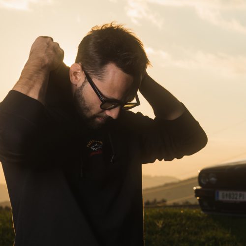A CD cover of a 35-year-old man with glasses trying to pull on the hood of his hoodie in the sunset.