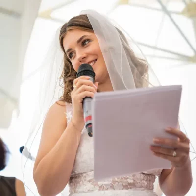 The bride gives her wedding speech. In one hand she holds a microphone, in the other a large sheet of paper. She smiles charmingly as she does so. In the background, a sea of white parasols.